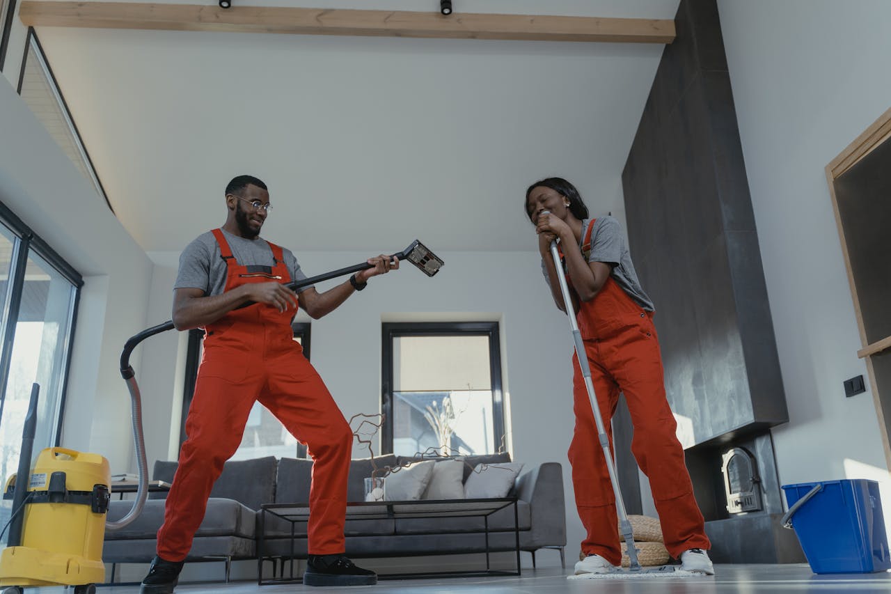 Two adults in red coveralls having fun while cleaning at home, using vacuum and mop.
